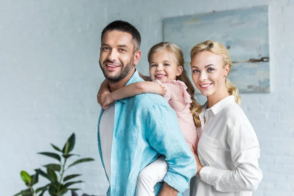 Genitori felici con adorabile figlioletta sorridente alla macchina fotografica mentre trascorrono del tempo insieme a casa — Foto stock