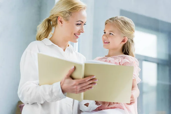 Glückliche Mutter und Tochter lächeln einander an, während sie zu Hause gemeinsam Bücher lesen — Stockfoto