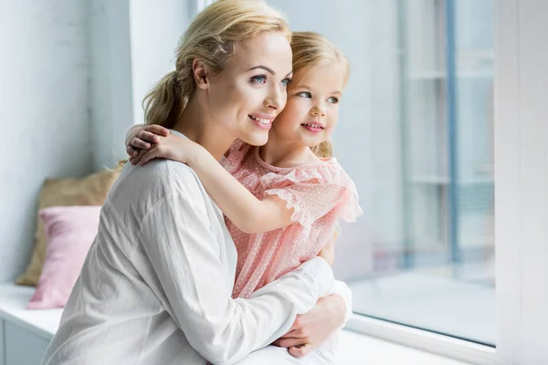 Hermosa madre feliz y la hija abrazando y mirando a la ventana - foto de stock