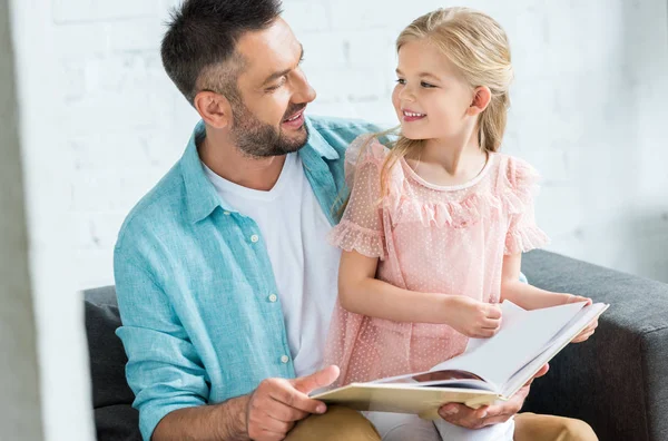Счастливые отец и дочь улыбаются друг другу во время чтения книги вместе дома — стоковое фото
