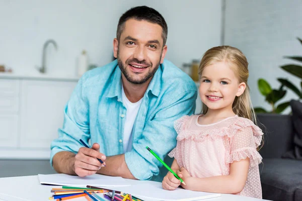 Feliz padre e hija dibujo con lápices de colores y sonriendo a la cámara - foto de stock