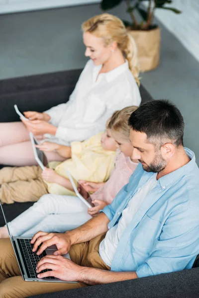 Visão de alto ângulo da família com duas crianças sentadas no sofá e usando dispositivos digitais em casa — Fotografia de Stock