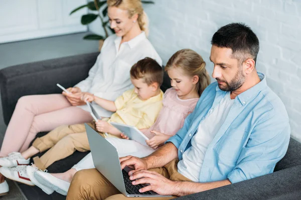 Сім'я з двома дітьми сидить на дивані і використовує цифрові пристрої вдома — стокове фото