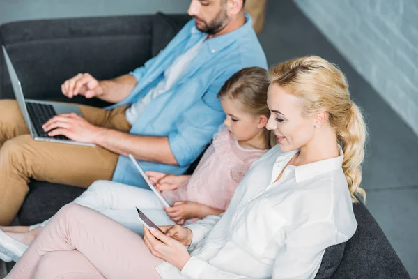 Visão de alto ângulo dos pais com a filha pequena bonito sentado no sofá e usando dispositivos digitais — Fotografia de Stock