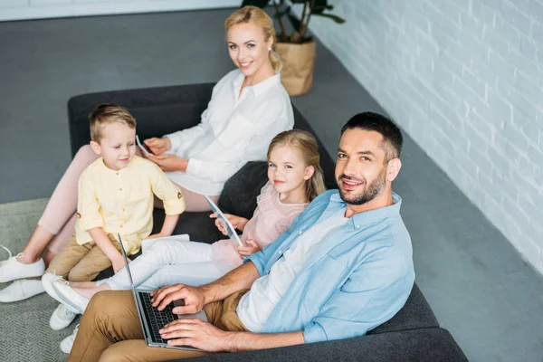 Vue grand angle de la famille heureuse en utilisant des appareils numériques et souriant à la caméra — Photo de stock