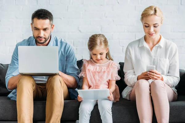 Padres y linda hija pequeña sentado en el sofá y el uso de dispositivos digitales - foto de stock