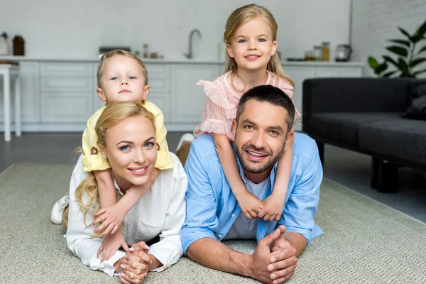 Genitori felici con due adorabili bambini sdraiati sul tappeto e sorridenti alla macchina fotografica a casa — Foto stock