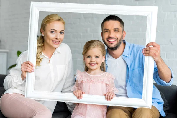 Happy parents with adorable little daughter holding white frame and smiling at camera — Stock Photo