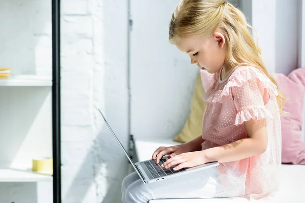 Vista lateral de un niño pequeño lindo usando el ordenador portátil mientras está sentado en el alféizar de la ventana - foto de stock