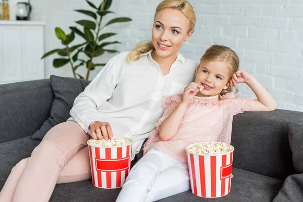 Beautiful smiling mother and daughter eating popcorn and looking away at home — Stock Photo