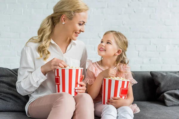 Mãe feliz e filha sorrindo uns aos outros enquanto comem pipocas em casa — Fotografia de Stock