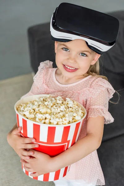 Красива дитина в гарнітурі віртуальної реальності тримає коробку з попкорном і посміхається на камеру — стокове фото