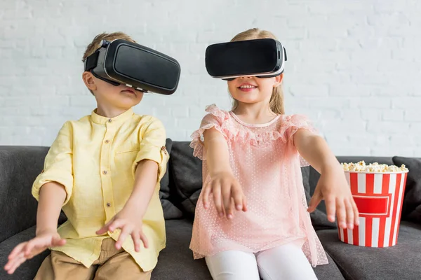 Entzückende kleine Kinder, die Virtual-Reality-Headsets benutzen, während sie zu Hause auf dem Sofa sitzen — Stockfoto