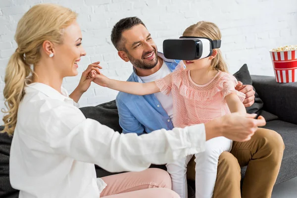 Pais felizes olhando para a filhinha bonito usando fone de ouvido realidade virtual em casa — Fotografia de Stock