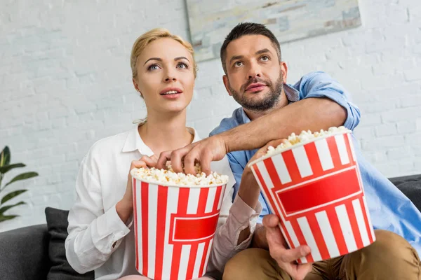 Low angle view of couple eating popcorn and looking away while sitting together at home — Stock Photo