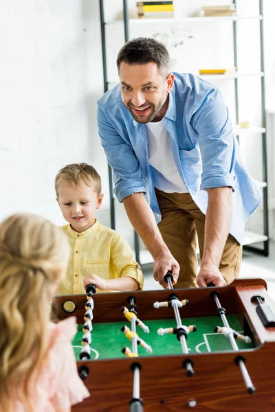 Счастливый отец с двумя очаровательными детьми, играющими в настольный футбол дома — стоковое фото
