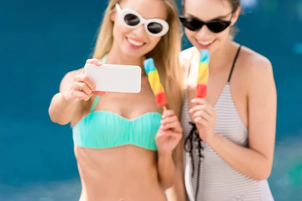 Mujeres jóvenes atractivas en traje de baño y bikini tomando selfie con paletas en la piscina - foto de stock