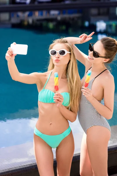 Hermosas mujeres jóvenes en traje de baño y bikini tomando selfie con paletas en la piscina - foto de stock