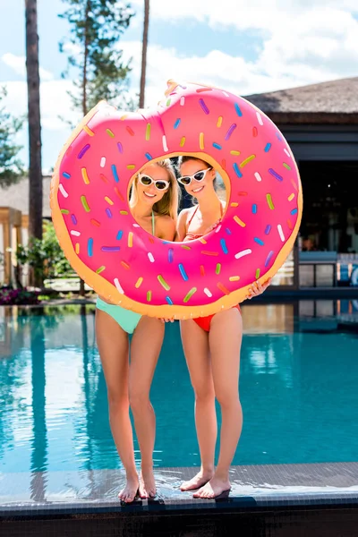 Sonrientes mujeres jóvenes mirando a la cámara a través de un anillo inflable en forma de rosquilla mordida en la piscina - foto de stock