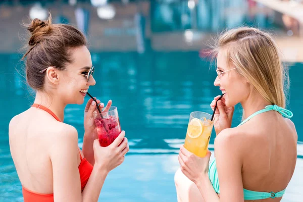 Hermosas mujeres jóvenes bebiendo deliciosas bebidas de frutas en la piscina y riendo - foto de stock