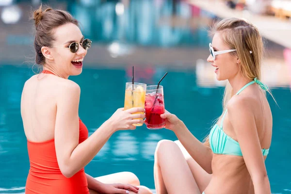 Vista lateral de atractivas mujeres jóvenes tintineo vasos de cócteles en la piscina - foto de stock