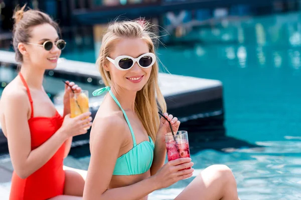 Mulheres jovens elegantes com deliciosas bebidas de frutas sentadas à beira da piscina — Fotografia de Stock
