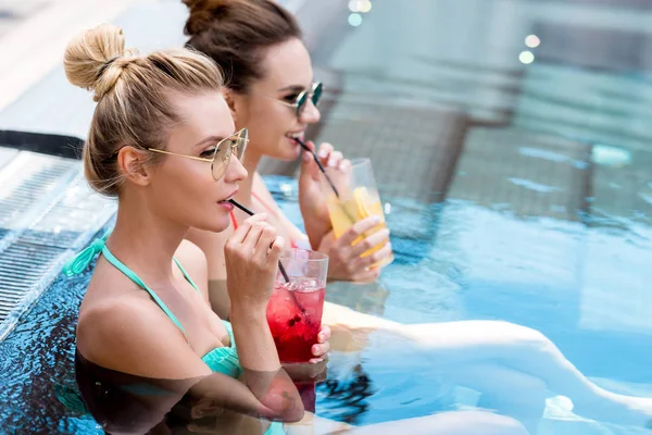 Hermosas mujeres jóvenes bebiendo deliciosos cócteles relajándose en la piscina y mirando al costado - foto de stock