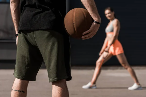 Selective focus of sportsman with basketball ball and sportswoman stretching on background — Stock Photo