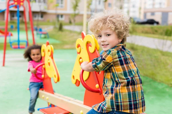 Selective focus of little smiling boy riding on rocking horse with curly child at playground — Stock Photo