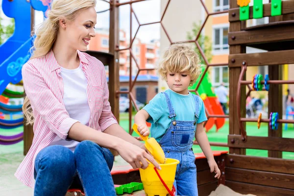 Entzückender kleiner Junge steckt Sand in Eimer mit Plastikschaufel, während seine Mutter Eimer im Sandkasten auf dem Spielplatz hält — Stockfoto