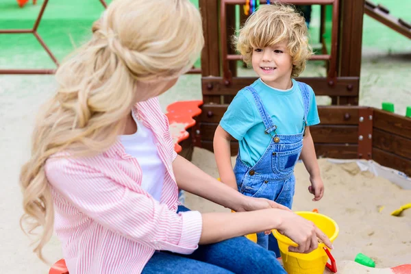 Visão traseira da mãe segurando balde de plástico e falando com o pequeno filho sorridente na caixa de areia no parque infantil — Fotografia de Stock
