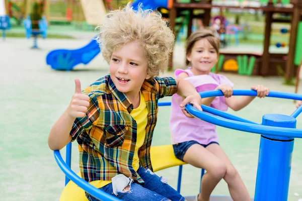 Adorável curly menino fazendo polegar até gesto enquanto montando no carrossel com criança pequena no playground — Fotografia de Stock