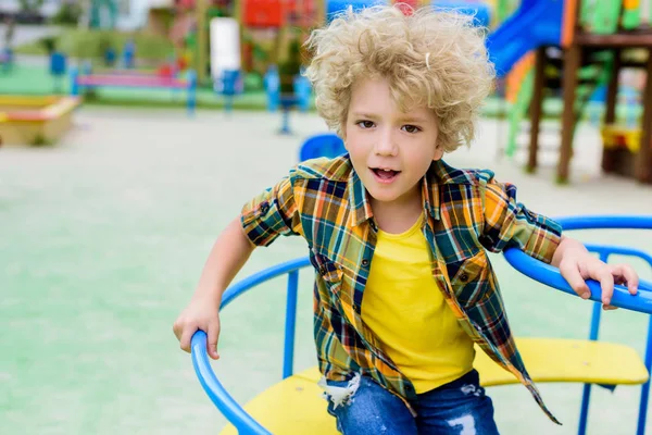 Selective focus of curly little boy riding on carousel at playground — Stock Photo