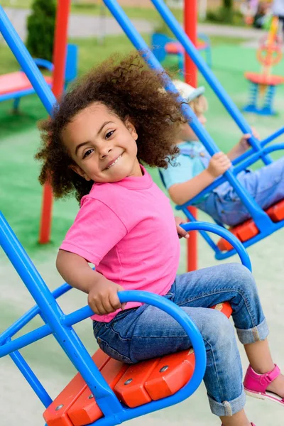 Sonriente rizado africano americano pequeño niño cabalgando en swing en patio de recreo - foto de stock