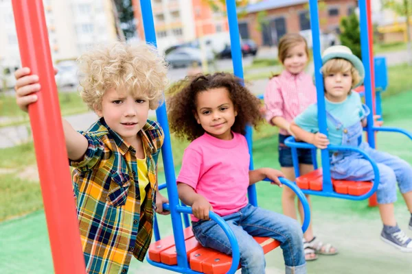 Grupo multicultural de niños pequeños montando en columpios en el patio de recreo - foto de stock