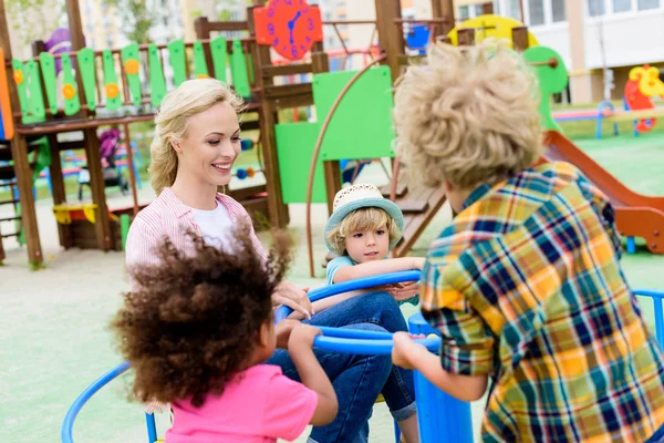 Happy mother riding with two sons and their african american friend on carousel at playground — Stock Photo