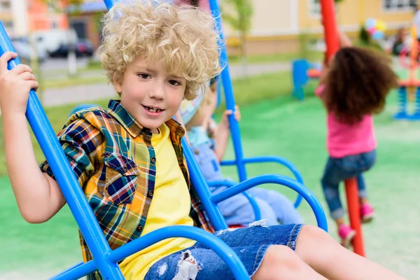 Селективный фокус счастливого кудрявого мальчика, катающегося на качелях на детской площадке — стоковое фото
