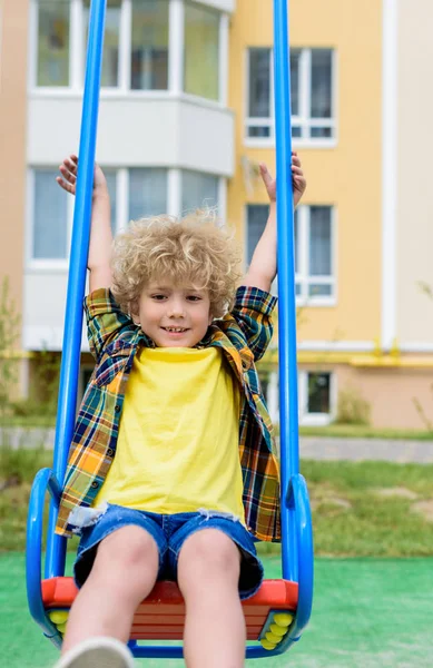 Niño rizado feliz montar en columpio en el patio de recreo - foto de stock