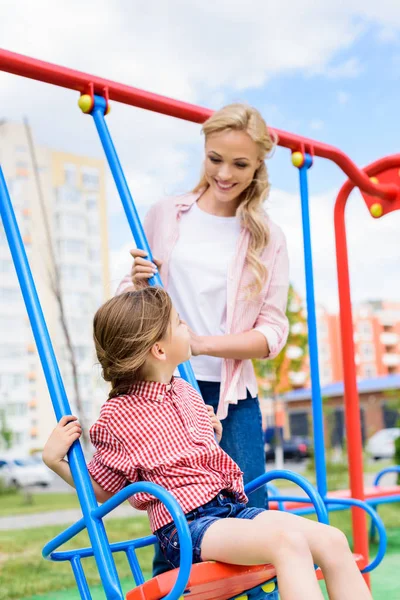 Selektiver Fokus der lächelnden Mutter, die Tochter auf Schaukel auf Spielplatz reitet — Stockfoto