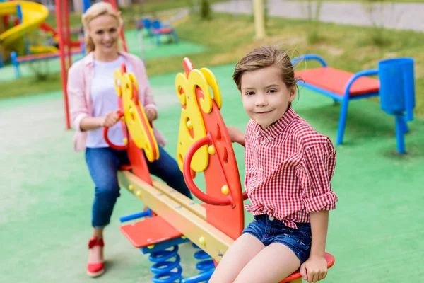 Selektiver Fokus eines entzückenden kleinen Kindes, das auf einem Schaukelpferd sitzt, während seine Mutter hinten auf dem Spielplatz sitzt — Stockfoto