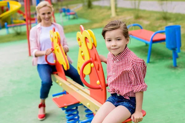 Messa a fuoco selettiva di adorabile bambino seduto sul cavallo a dondolo mentre sua madre seduta dietro al parco giochi — Foto stock