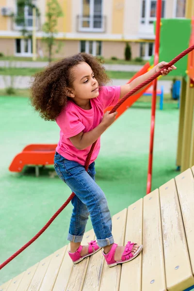 Vista lateral da criança americana africana encaracolada escalando com corda no playground — Fotografia de Stock