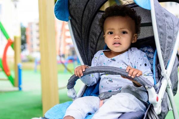 Selective focus of african american toddler sitting in baby carriage at playgorund — Stock Photo