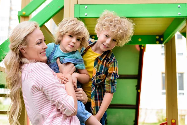 Vista lateral de la madre sonriente con dos adorables hijos pequeños en el patio de recreo - foto de stock