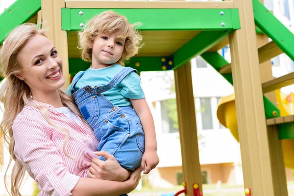 Sonriente madre sosteniendo adorable pequeño hijo en las manos en el patio de recreo - foto de stock