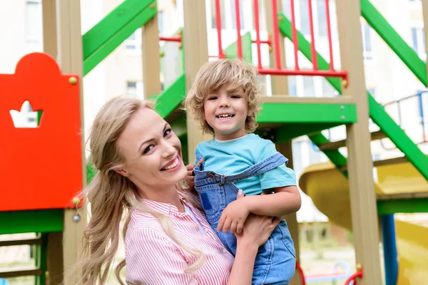 Retrato de madre feliz sosteniendo en las manos sonriente pequeño hijo en el patio de recreo - foto de stock
