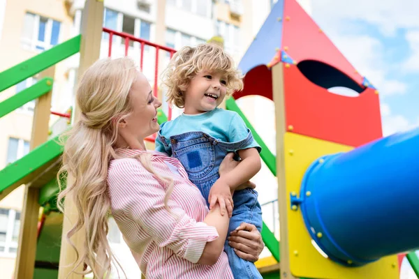 Vue à angle bas de mère heureuse tenant sur les mains souriant petit fils à aire de jeux — Photo de stock