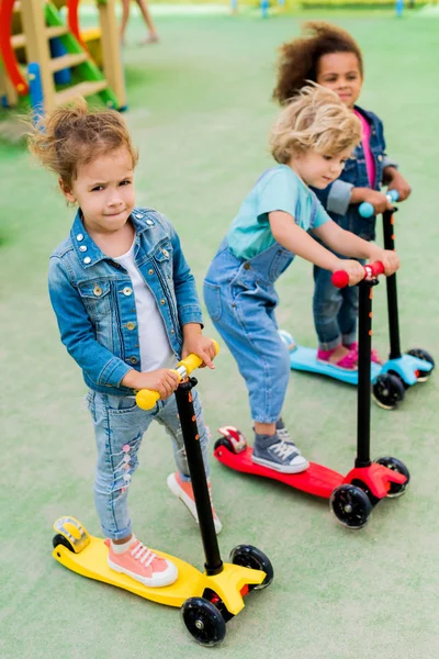 Focus selettivo dei bambini adorabili multiculturali che cavalcano sugli scooter a calci al parco giochi — Foto stock