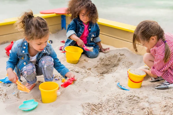 Tres niños pequeños multiétnicos jugando con cucharas de plástico y cubos en caja de arena en el patio de recreo - foto de stock