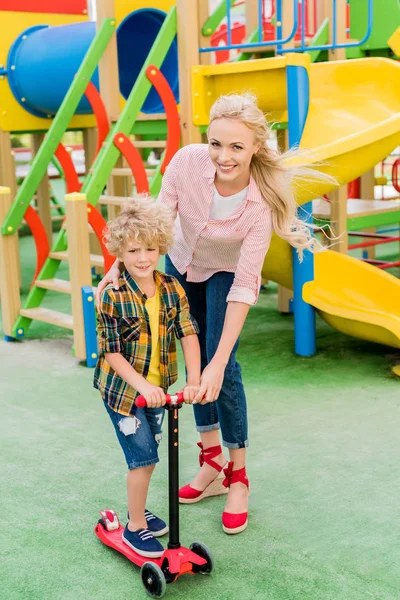 Mère souriante embrassant adorable fils bouclé alors qu'il roulait sur trottinette de coup de pied à l'aire de jeux — Photo de stock